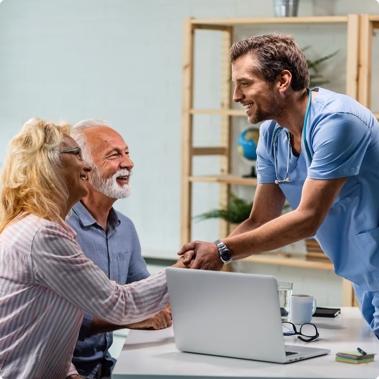 Smiling male doctor shaking hands with an elderly woman while an elderly man looks on in a medical office.