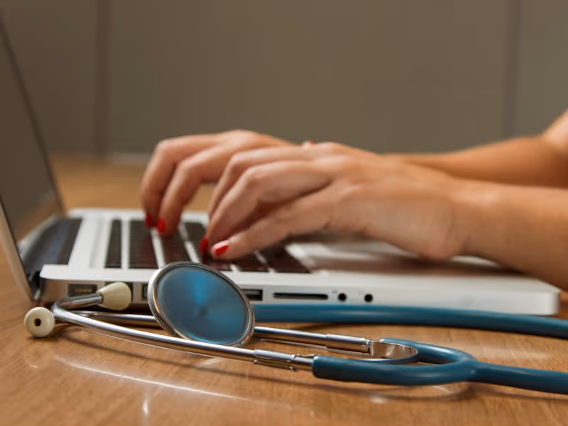 Person typing on a laptop keyboard with a blue stethoscope placed on a wooden table nearby.