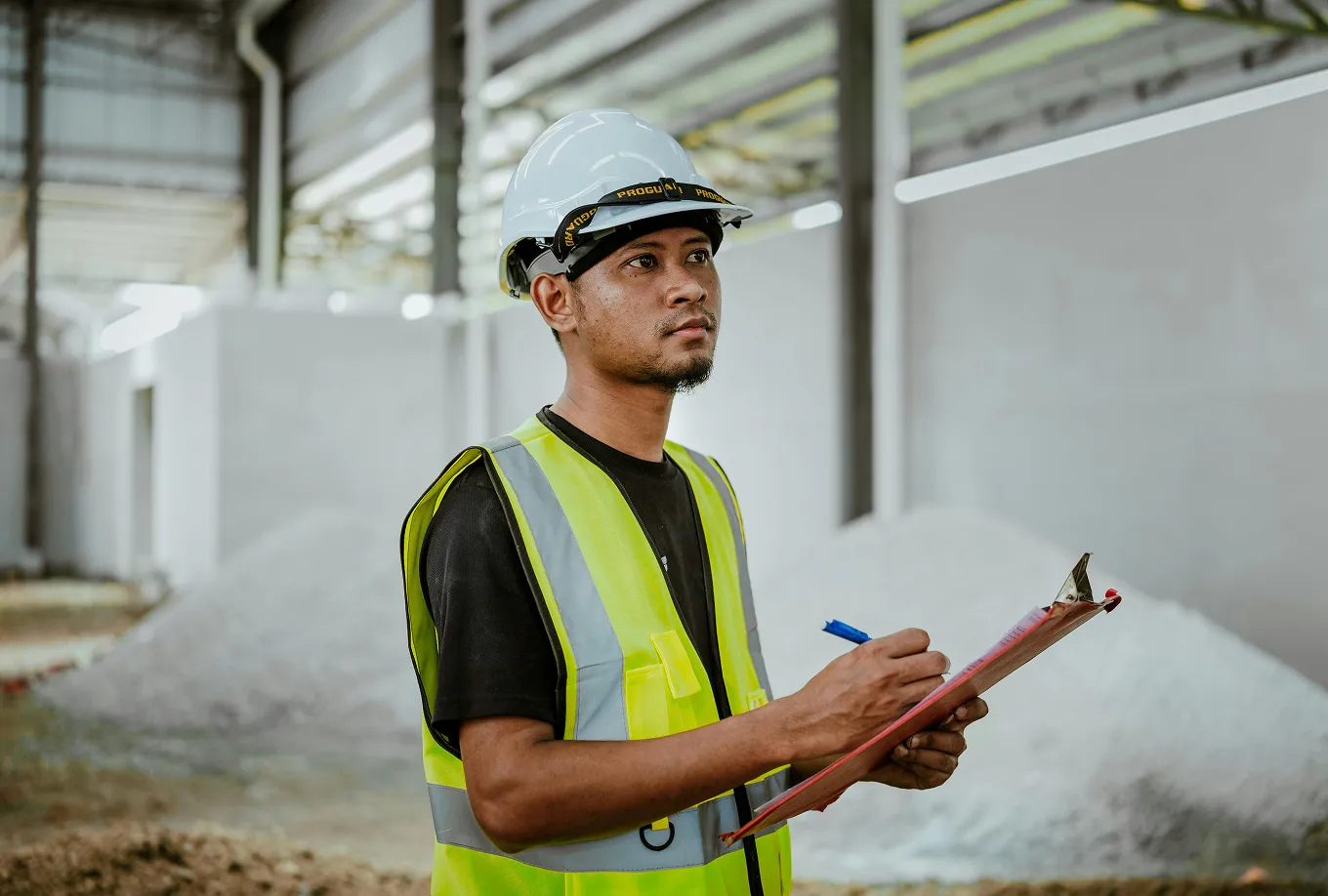 Construction worker wearing a white hard hat and yellow safety vest writing on a clipboard inside a building under construction.