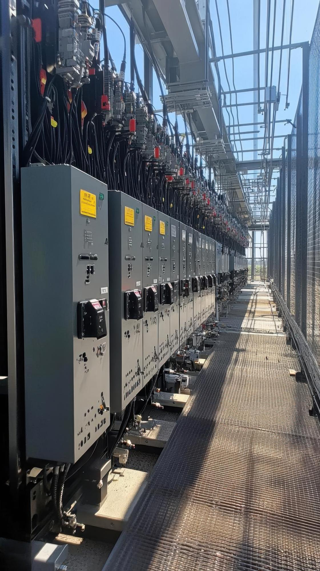 Row of industrial electrical control panels with numerous cables and switches inside a metal-enclosed facility with a grated floor.