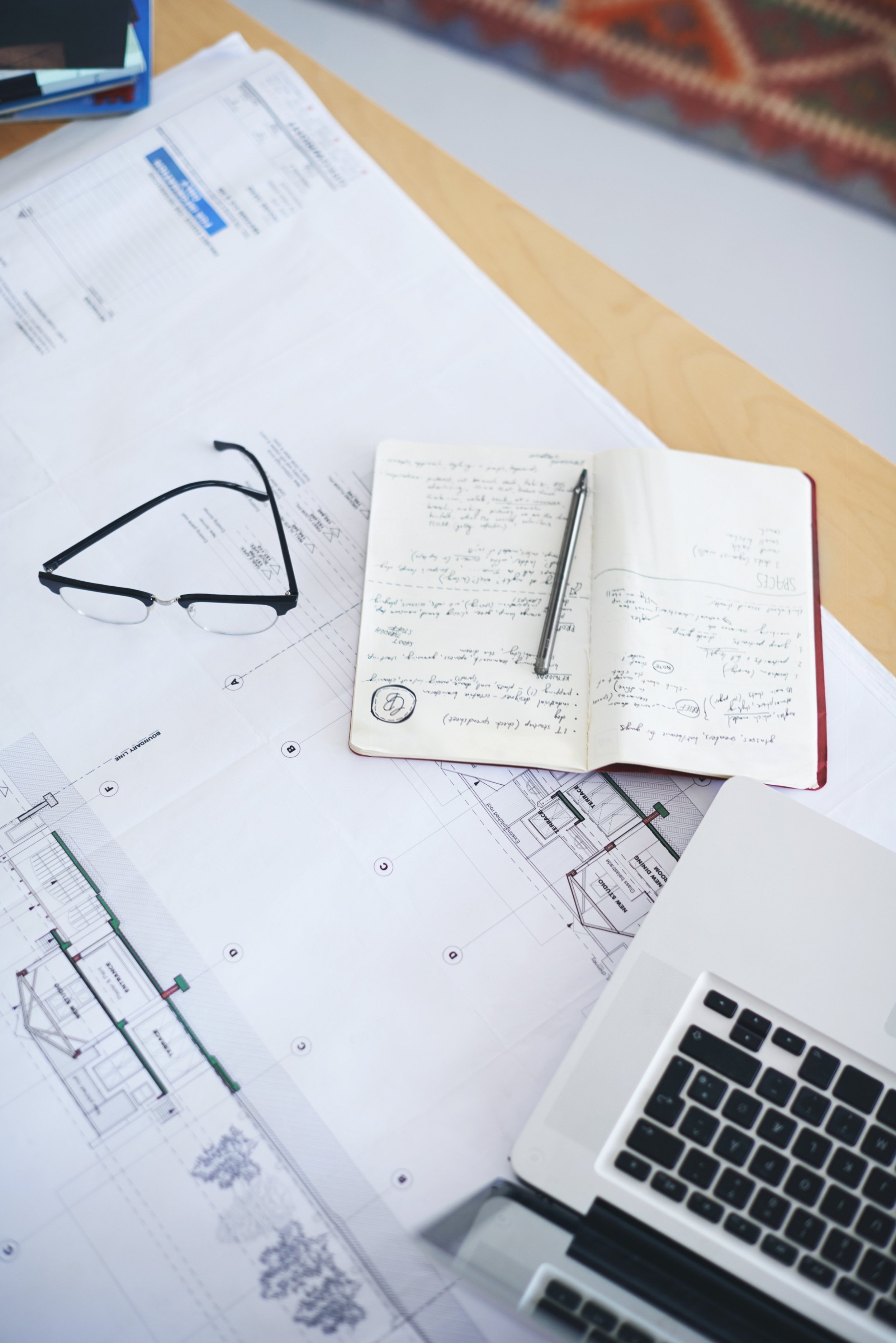 Desk with architectural blueprints, open notebook with handwritten notes and pen, black eyeglasses, and silver laptop keyboard.