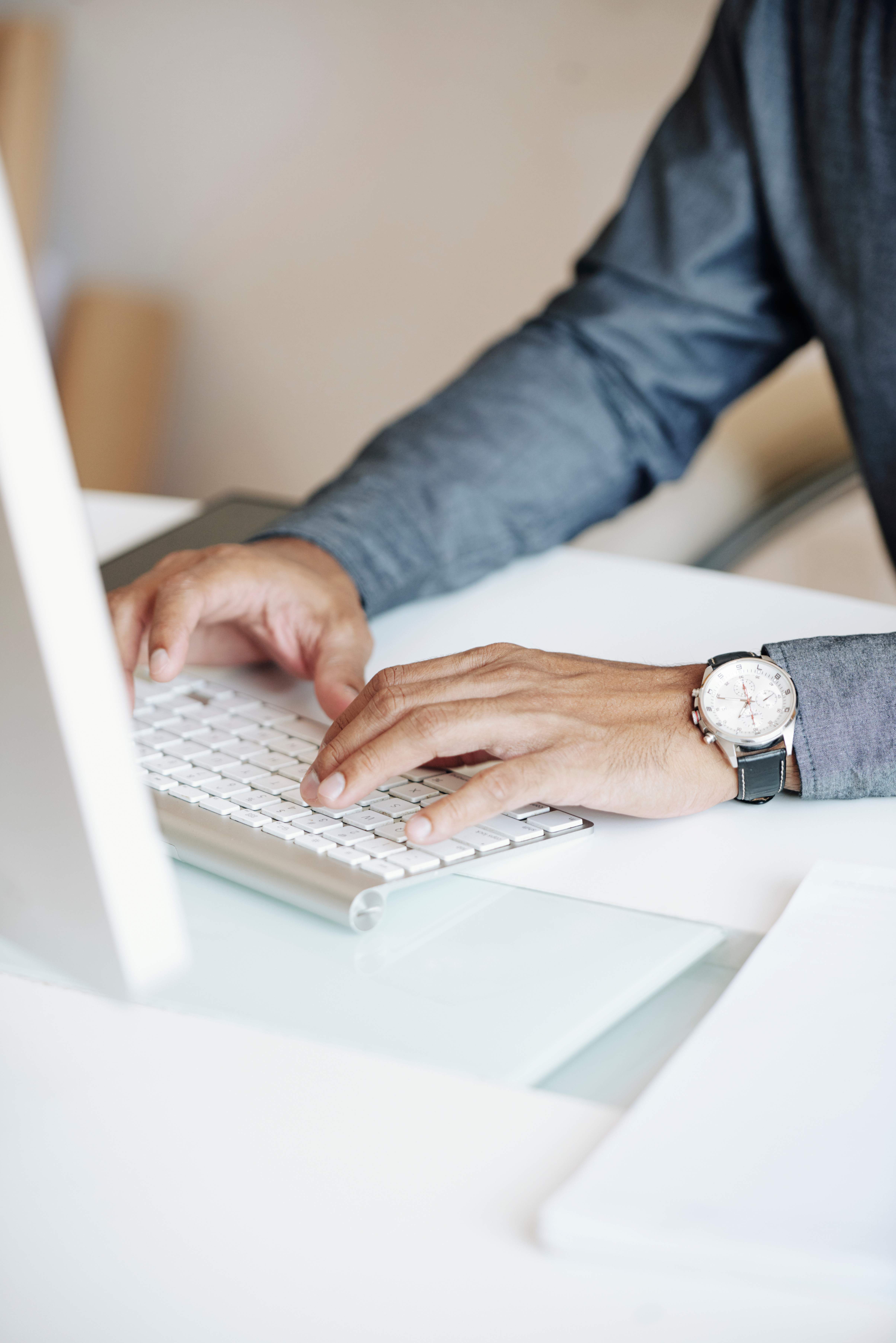 Person typing on a white keyboard next to a computer monitor, wearing a watch and a long-sleeve dark shirt.