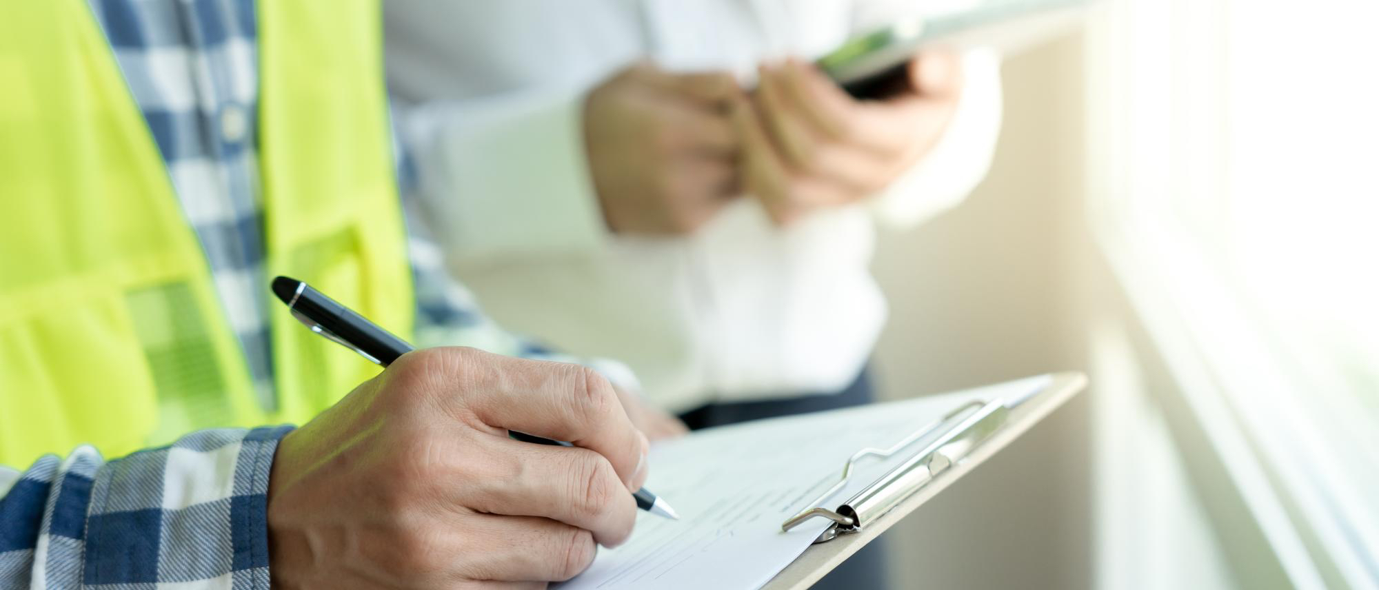 Close-up of a person in a yellow safety vest writing on a clipboard with another person holding a tablet in the background.