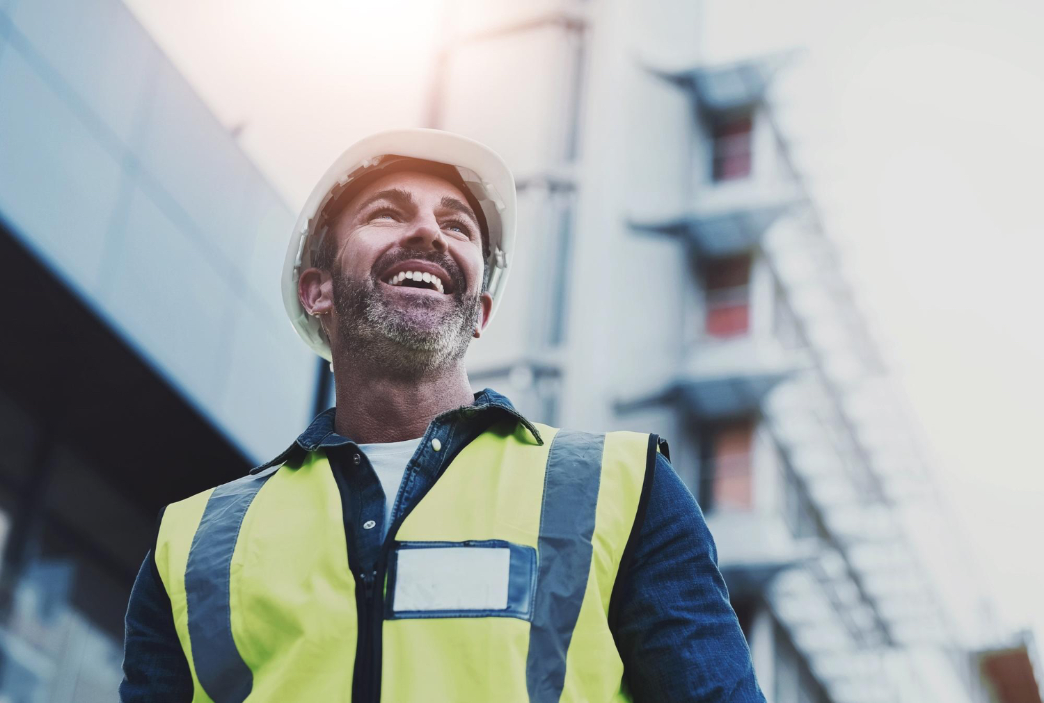 Smiling construction worker wearing a white hard hat and yellow safety vest at a building site.