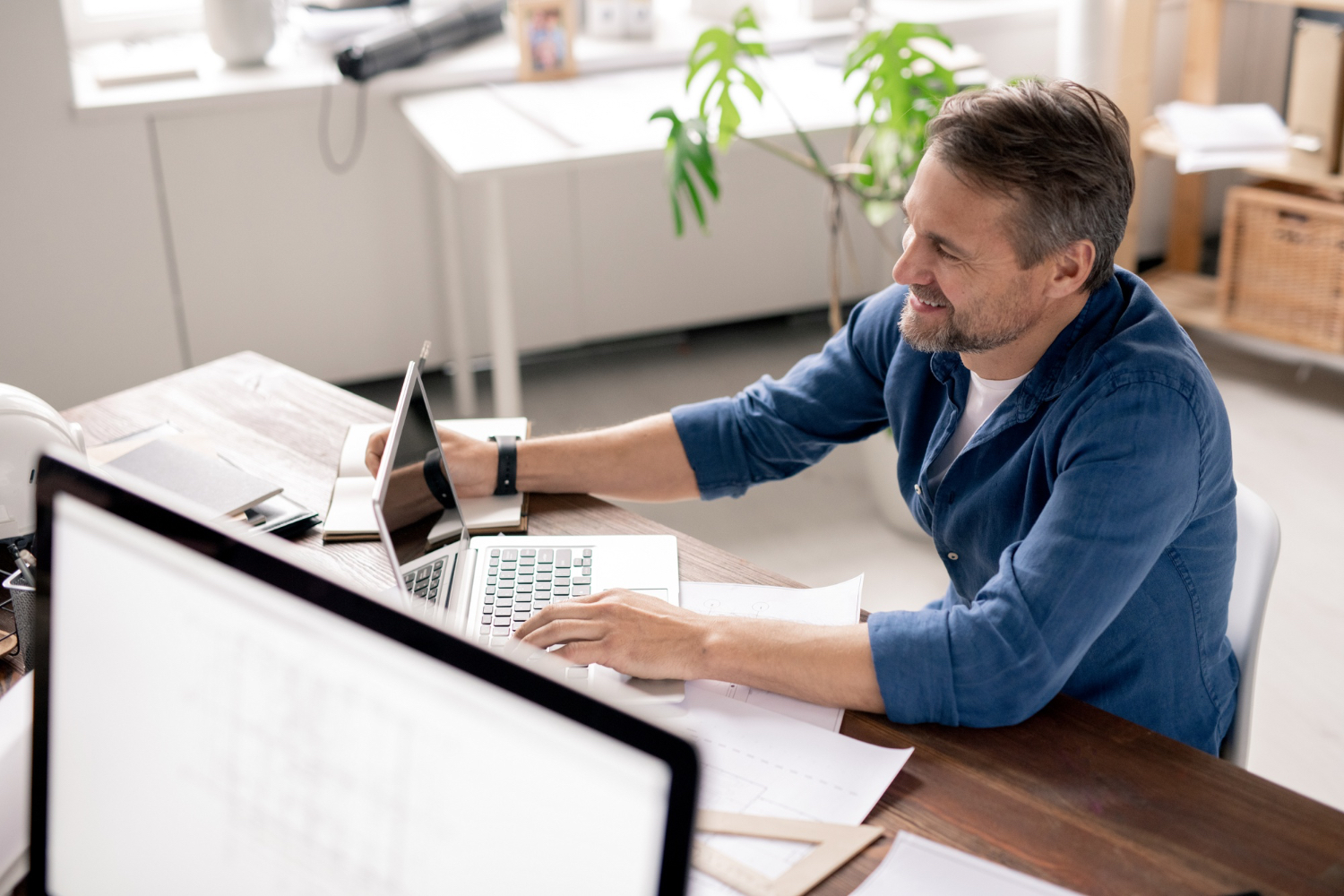 Smiling man in blue shirt working on a laptop at a desk with documents and a plant in the background.