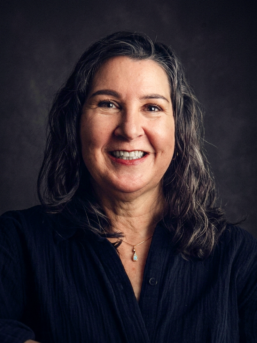 Smiling middle-aged woman with dark wavy hair wearing a black shirt and a pendant necklace against a dark background.