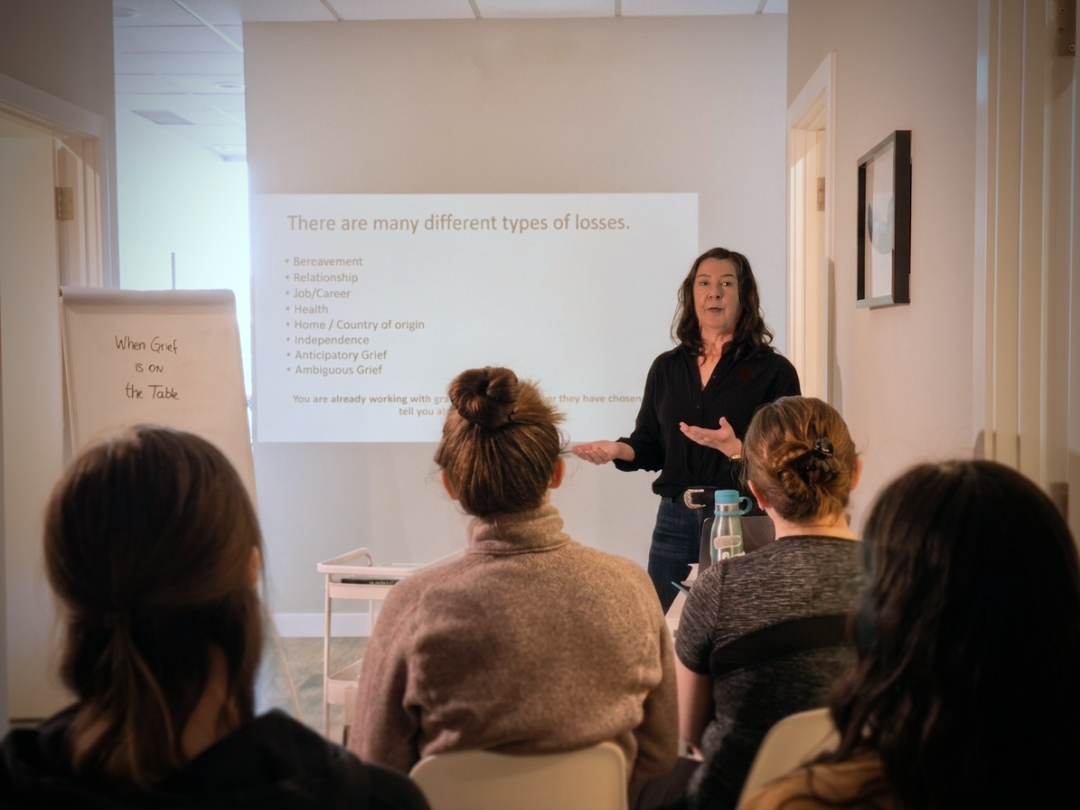 Woman giving a presentation to an audience on different types of losses, with a projection showing various kinds of grief and a flip chart with the text 'When Grief is on the Table'.
