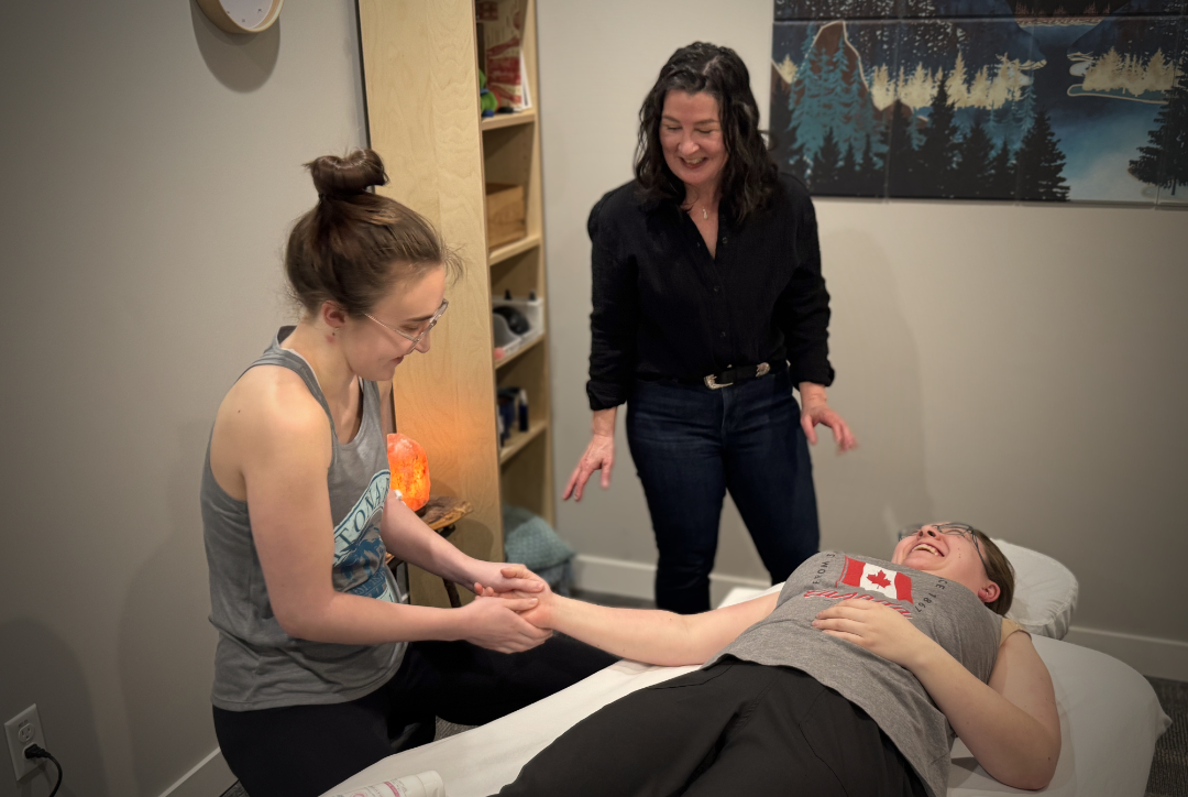 A massage therapist holding the hand of a smiling woman lying on a treatment table while another woman watches and smiles in a cozy clinic room.