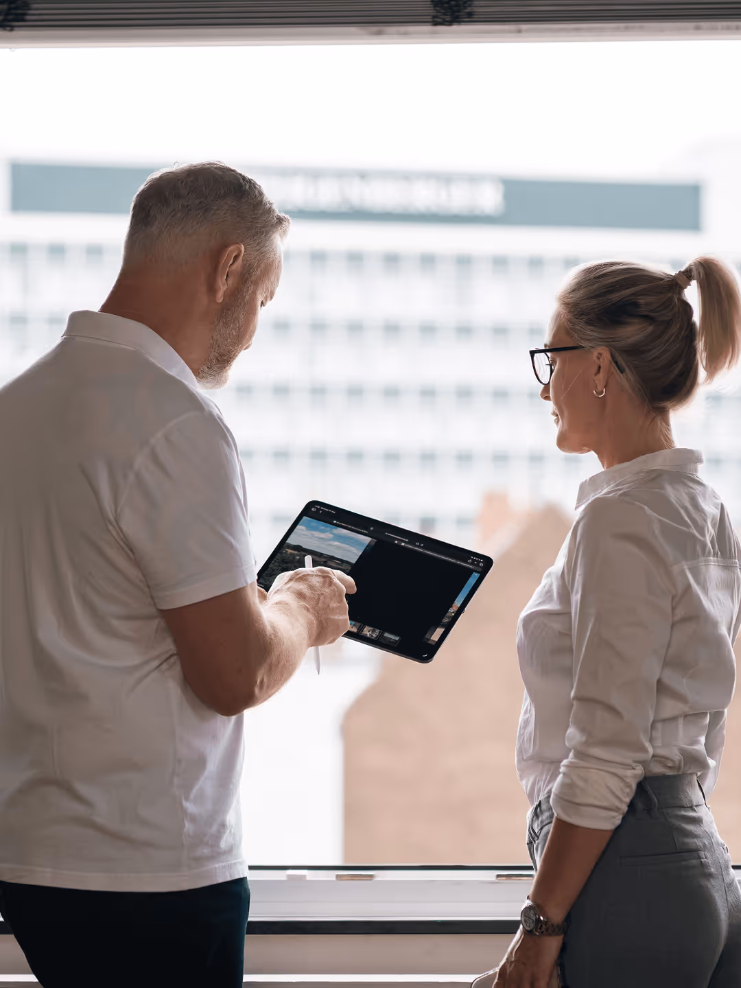 Zwei Personen betrachten ein Tablet vor einem Fenster, Gebäude im Hintergrund. Business-Kontext, Techniknutzung.