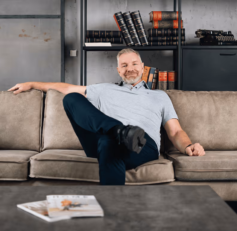 Mann entspannt auf grauem Sofa vor Bücherregal, Zeitschriften auf Couchtisch.