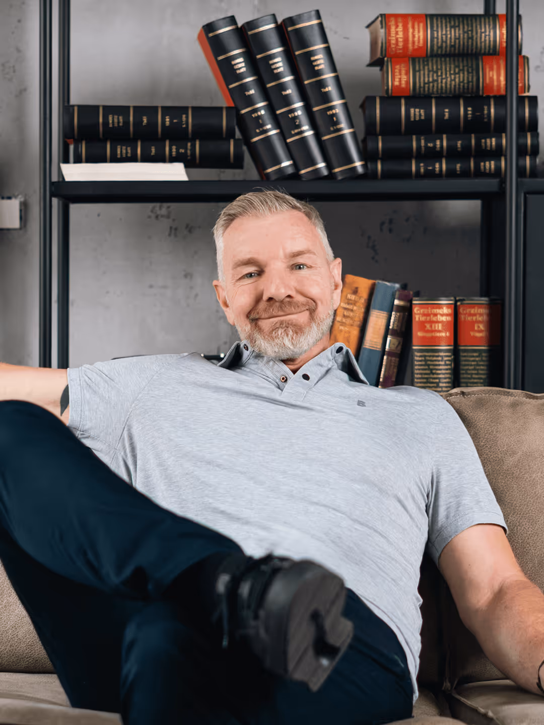 Lächelnder Mann auf Sofa, entspannt; Bücherregal im Hintergrund mit ledergebundenen Büchern; gemütliche Wohnzimmeratmosphäre.