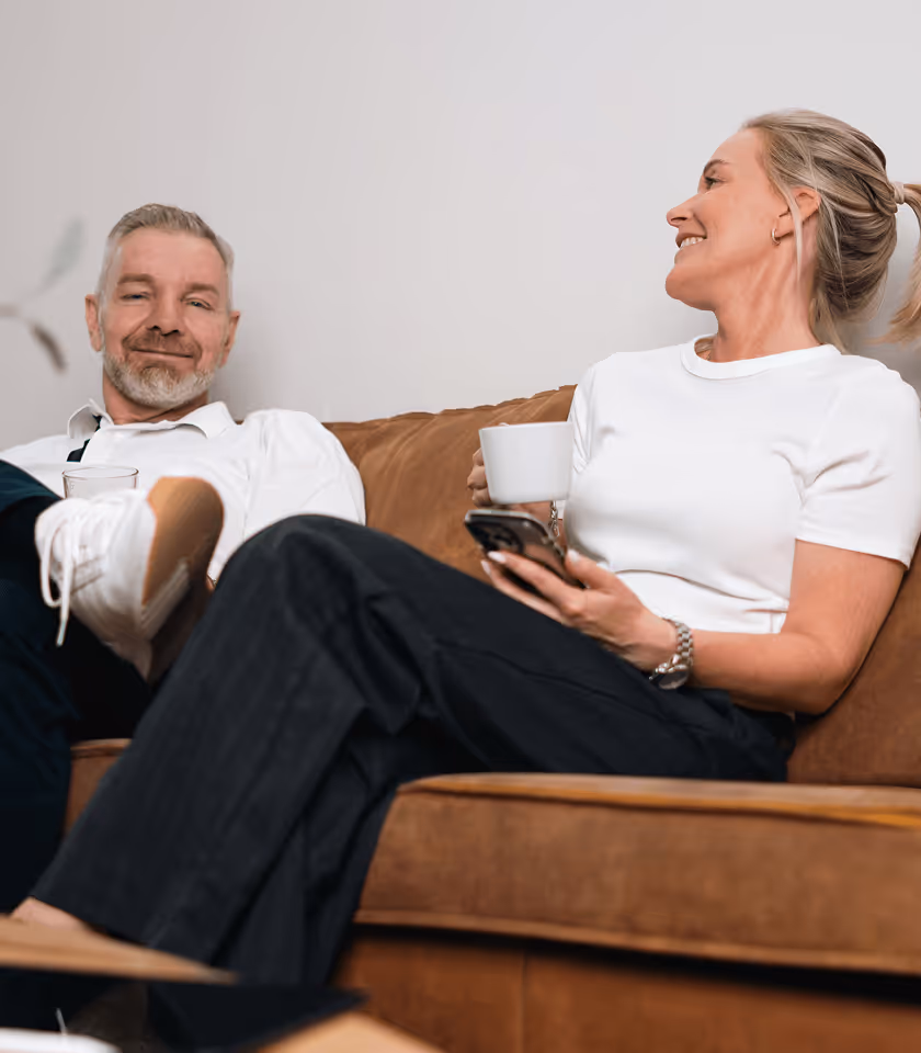 Älteres Paar lacht auf einem Sofa, sie hält eine Tasse und ein Smartphone, er trägt weiße Sneakers.