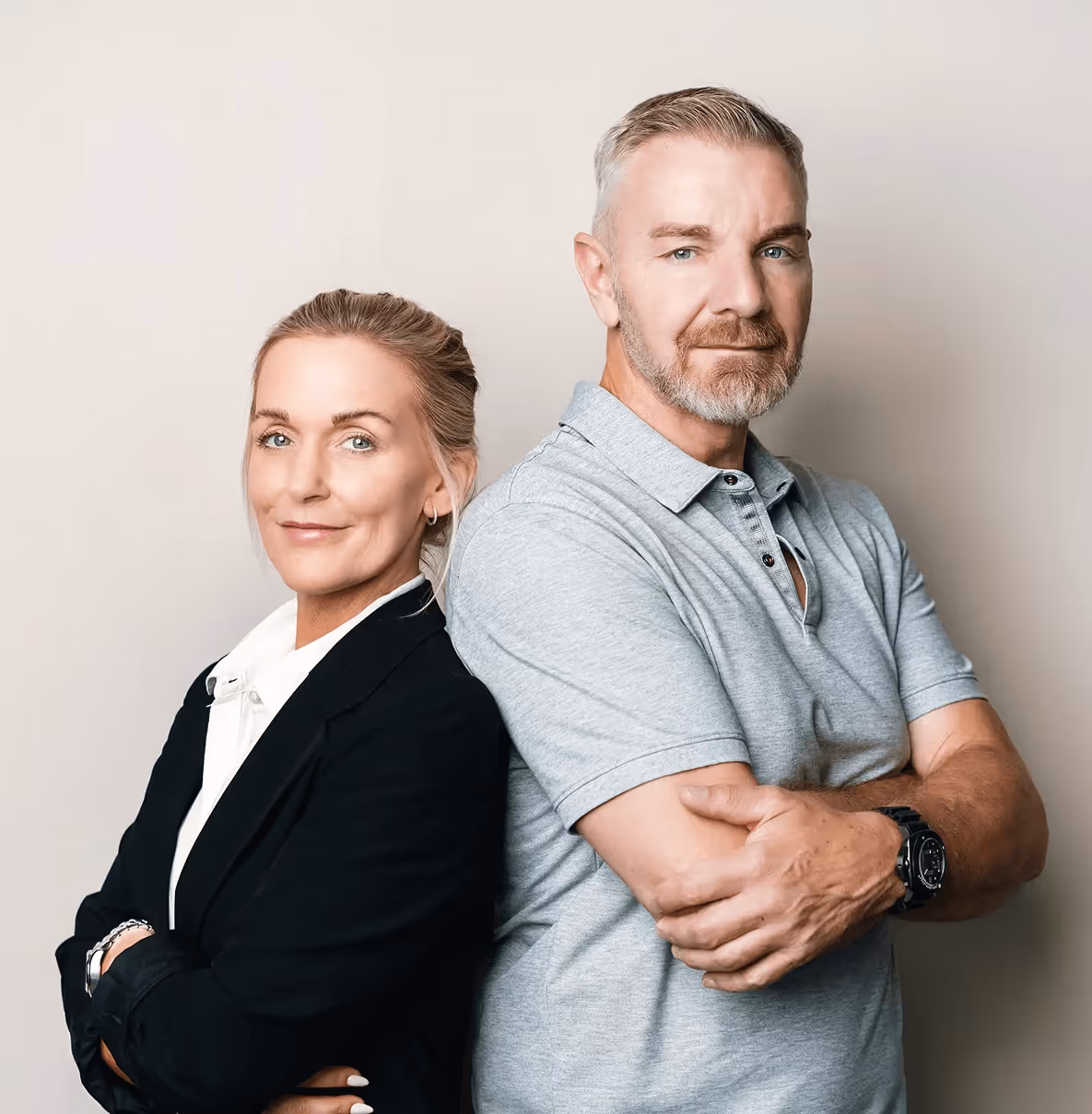Zwei Erwachsene stehen Rücken an Rücken vor neutralem Hintergrund, beide lächeln leicht, Frau in Anzugjacke, Mann in Polohemd.