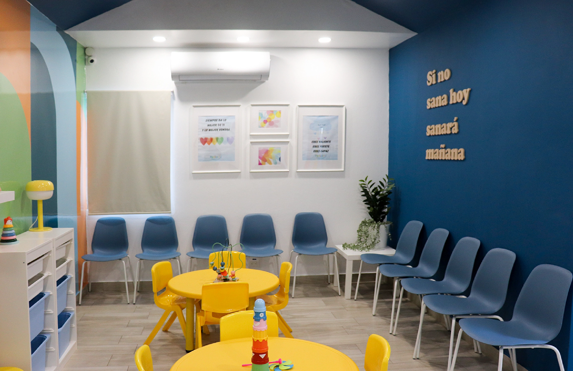 Children's waiting area with yellow tables and chairs, blue chairs along the walls, colorful wall art, and a blue wall with Spanish text.