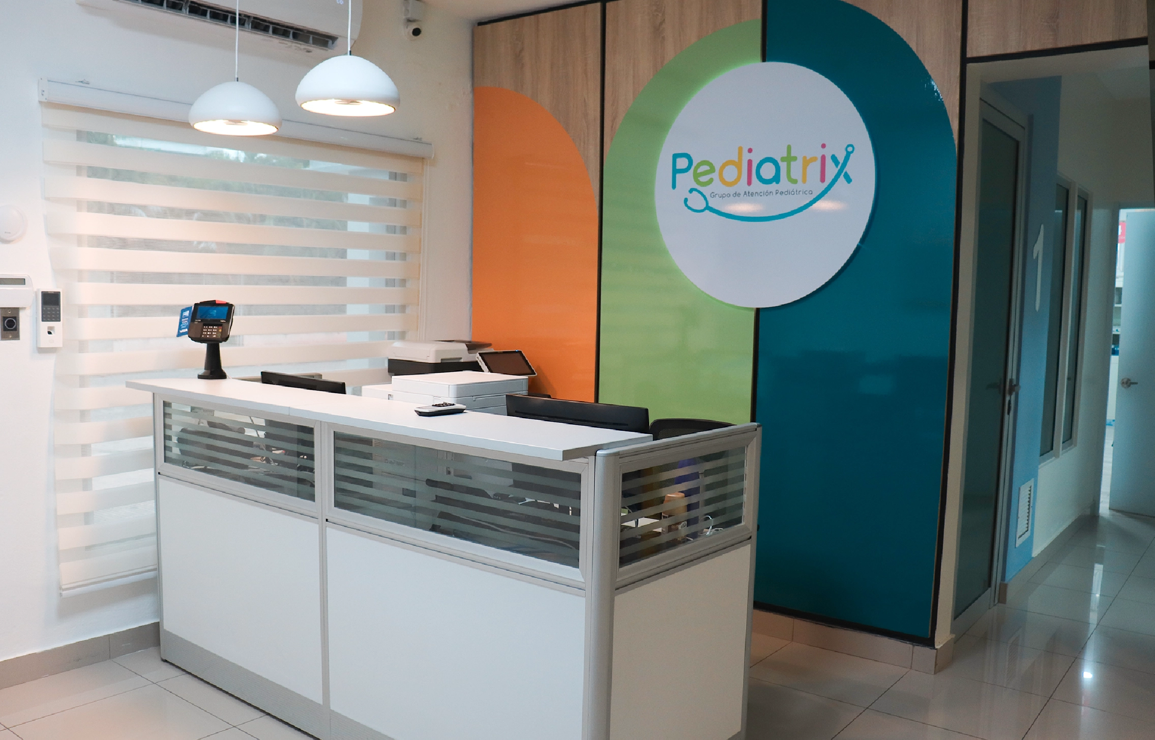 Reception area with white desk, two hanging lights, and colorful wall panel with Pediatrix logo in a pediatric clinic.