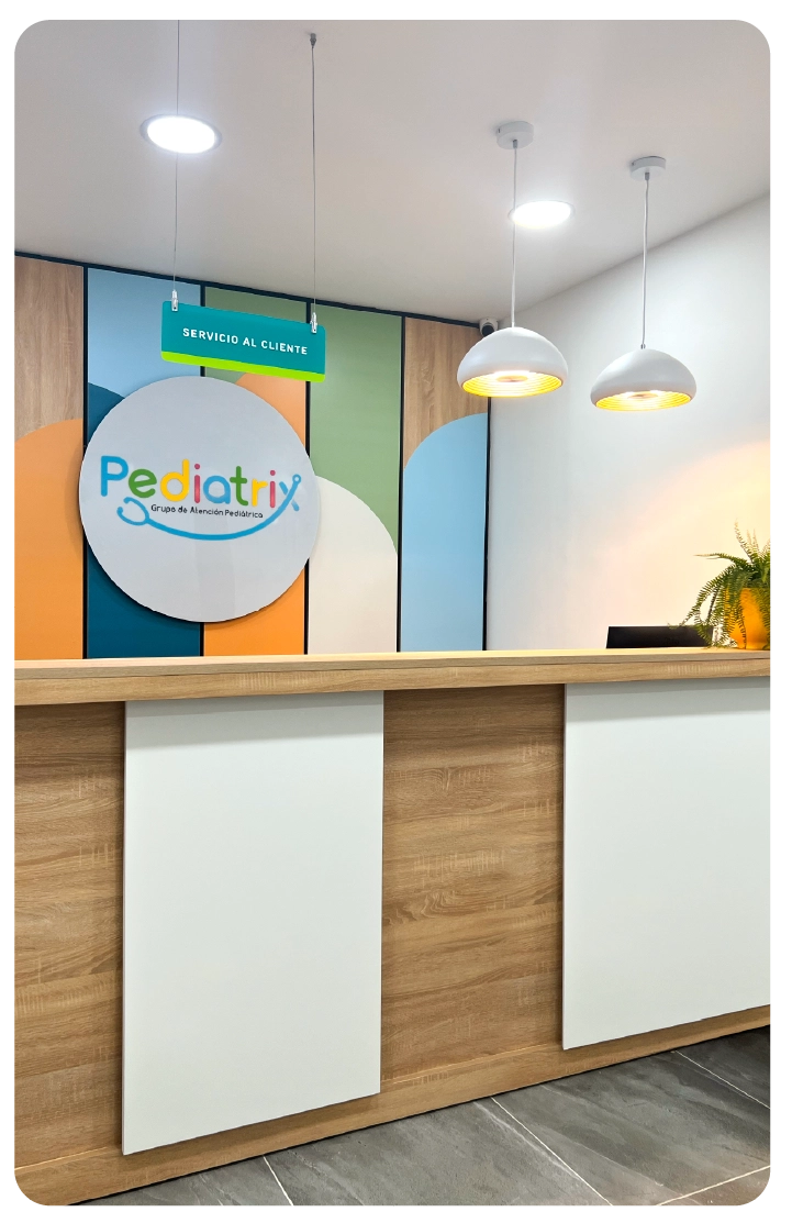 Pediatric clinic reception area with colorful wall paneling, a wooden desk, two hanging pendant lights, and a 'Servicio al Cliente' sign.