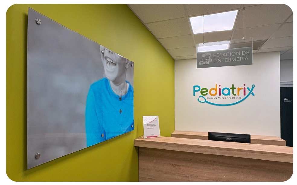 Reception desk in a pediatric care facility with a colorful 'Pediatrix Grupo de Atención Pediátrica' logo on a white wall and a blurred photo of a child on a green wall.