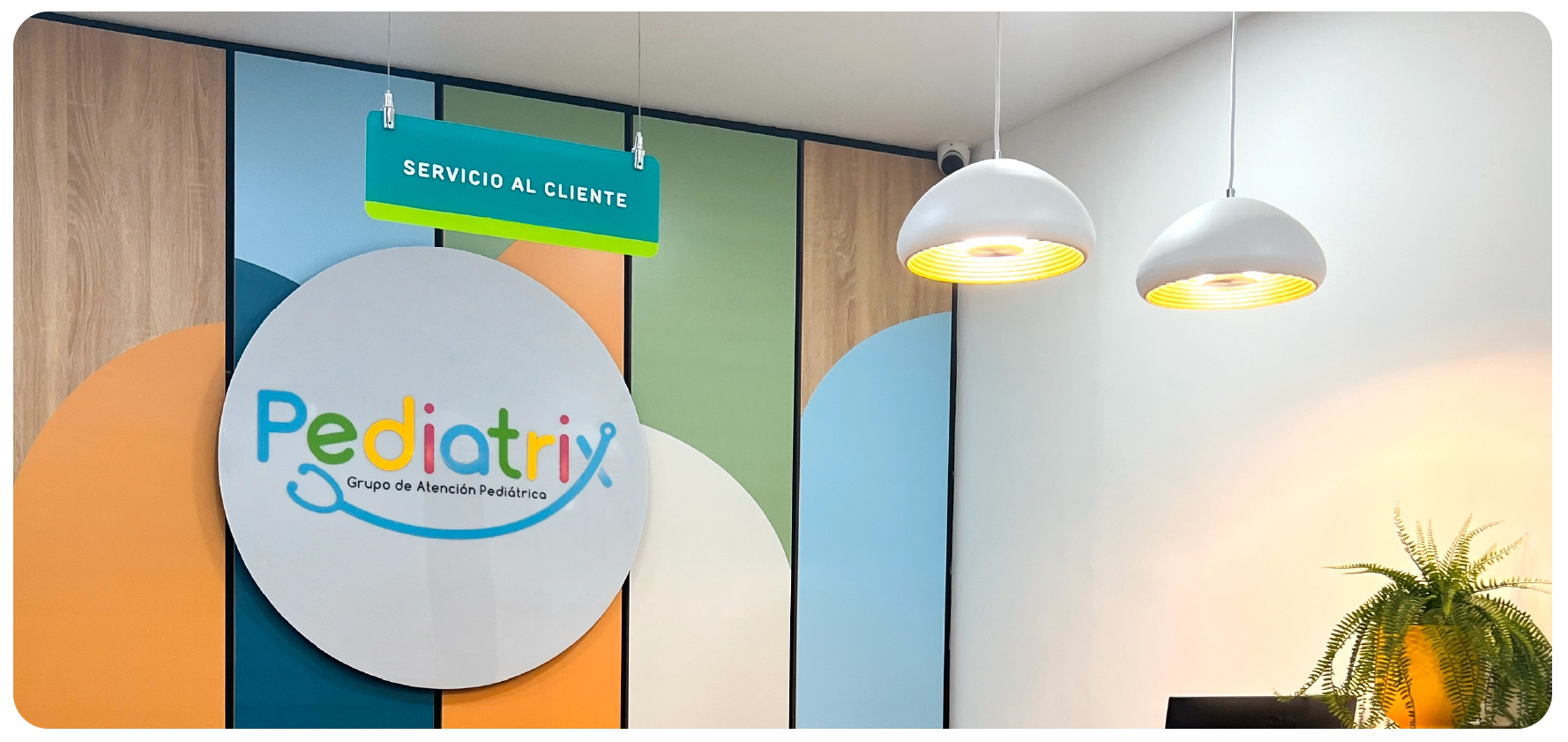 Colorful Pediatrix logo on a round sign with 'Grupo de Atención Pediátrica' below, against a multi-colored wall and two white hanging lights.