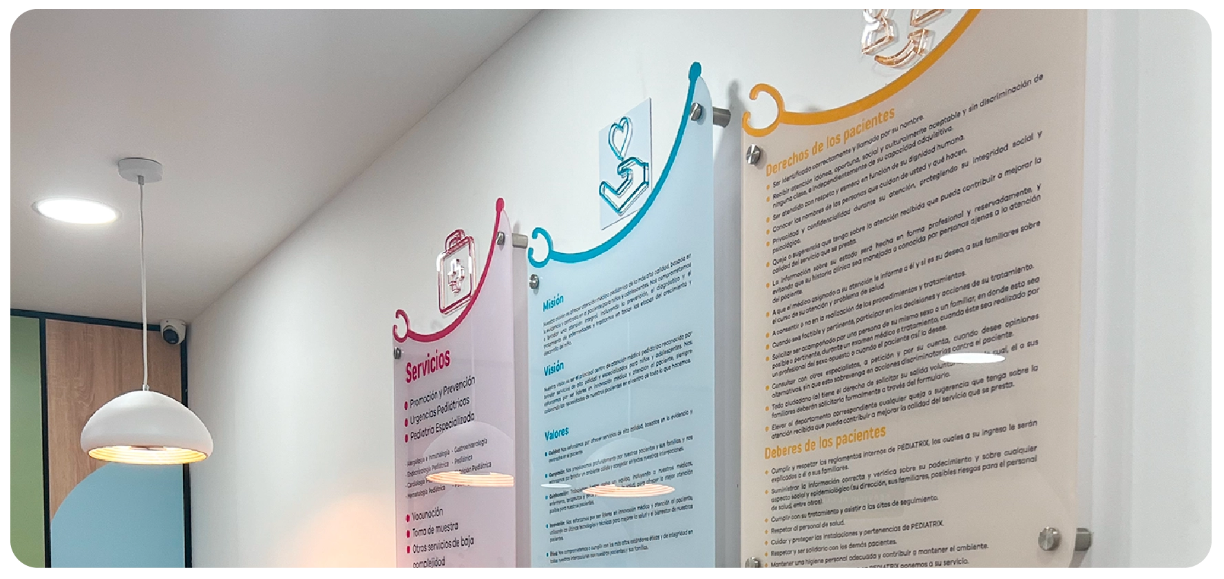 Wall-mounted medical information panels in Spanish with sections on services, mission, vision, values, patient rights, and patient duties, next to a hanging lamp and a security camera in a medical facility.