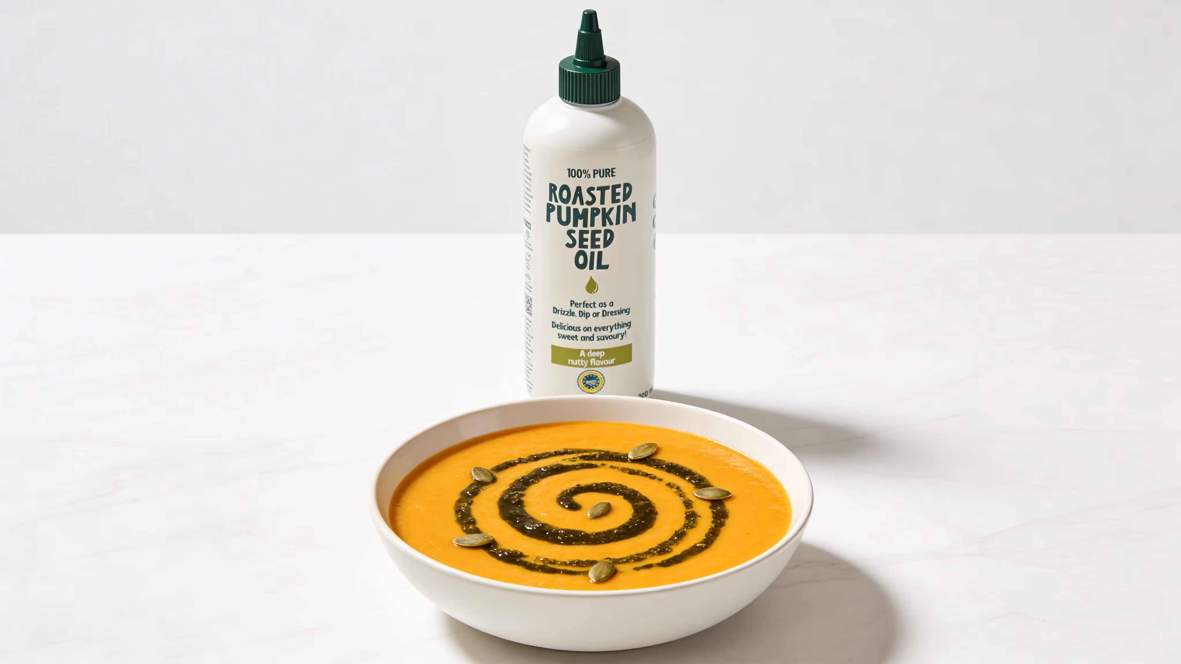 Bowl of creamy orange soup garnished with a green spiral drizzle and pumpkin seeds, with a bottle of roasted pumpkin seed oil behind it.