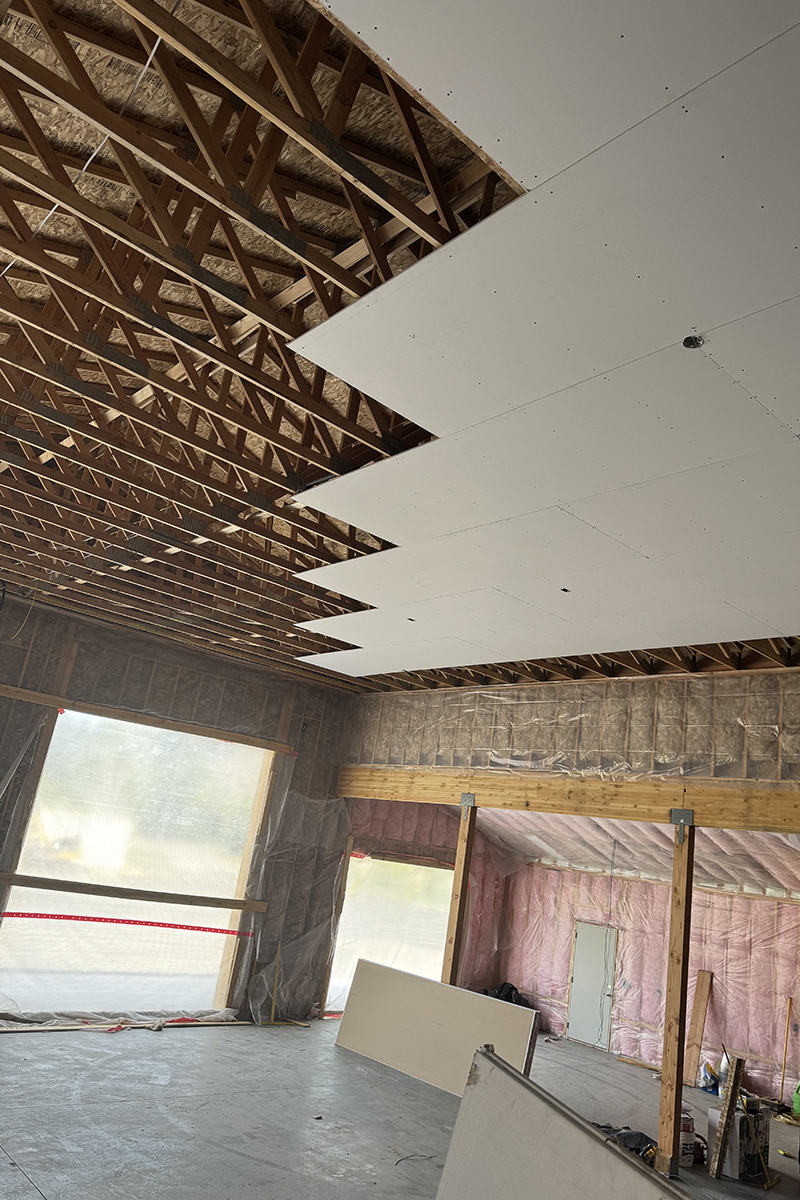 Interior of a building under construction showing exposed wooden ceiling trusses and partially installed drywall ceiling panels.