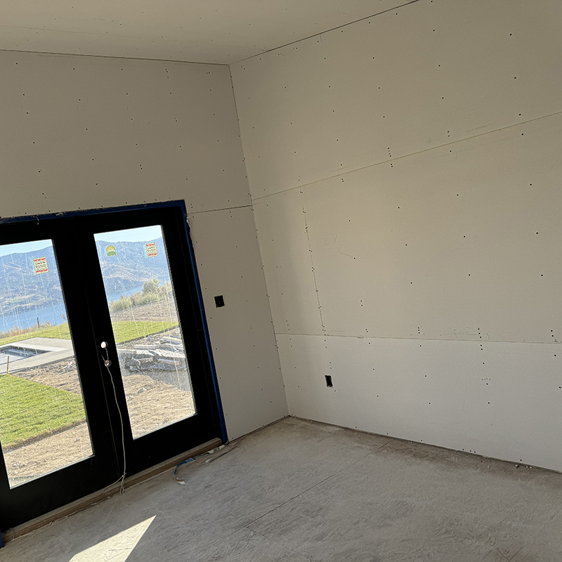 Unfinished interior room with drywall walls and concrete floor, featuring black-framed glass double doors opening to a grassy outdoor area with a pool and hills in the background.
