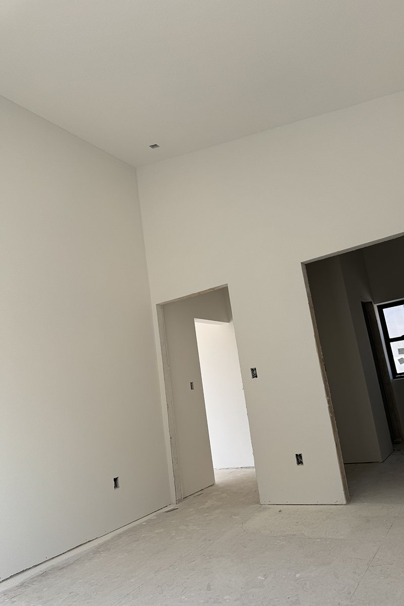 Empty room under construction with bare floors, white walls, and two open doorways leading to adjacent spaces.