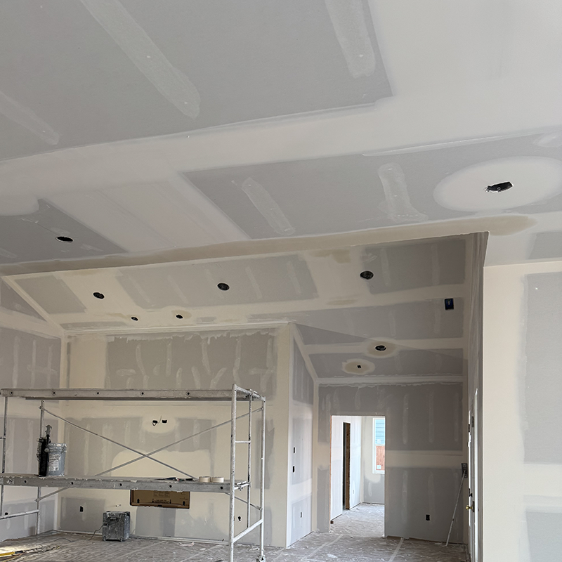 Interior room under construction with drywall installation, visible scaffolding, and unfinished ceiling with recessed light openings.