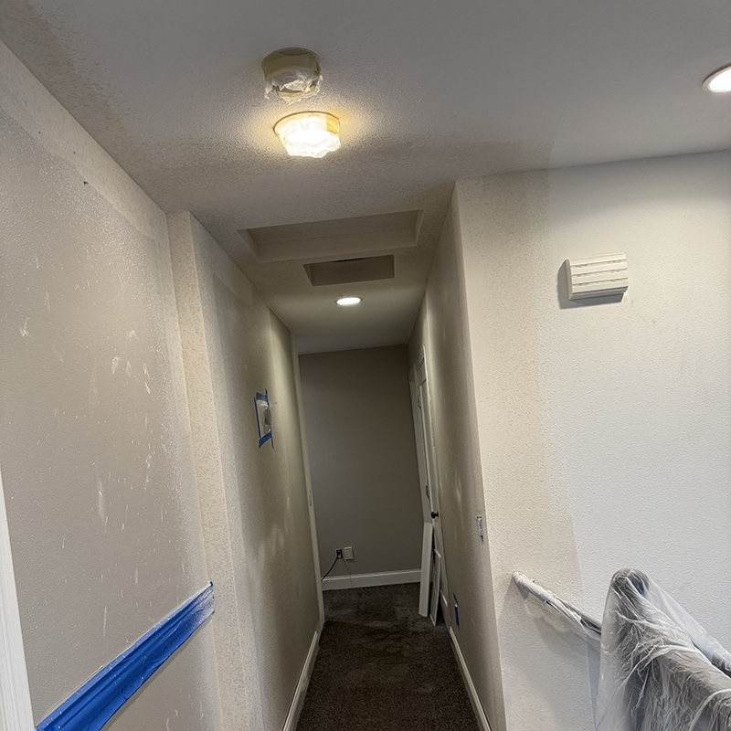 Dimly lit narrow hallway with beige walls and brown carpet undergoing renovation, with taped areas and a partially covered chair.