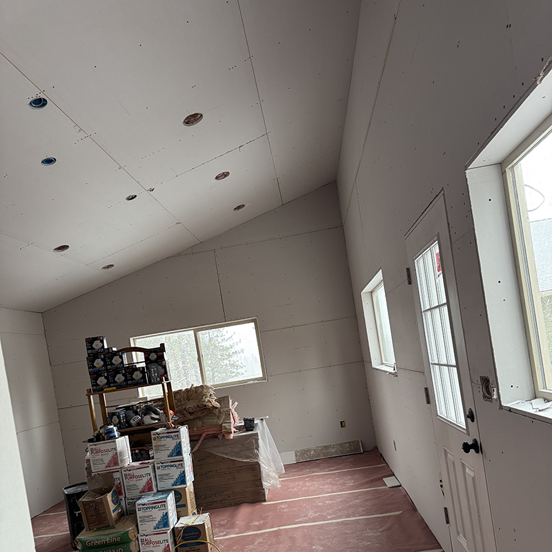 Interior of a room under construction with drywall installed, a door with windows, two other windows, and stacked boxes and insulation materials on a pink protective floor covering.