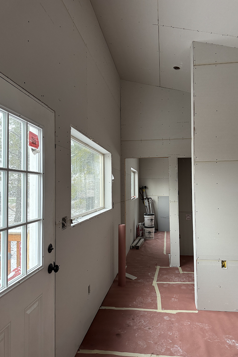 Interior of a room under construction with drywall installed, a door with glass panes on the left, a window, and construction materials on a red protective floor covering.