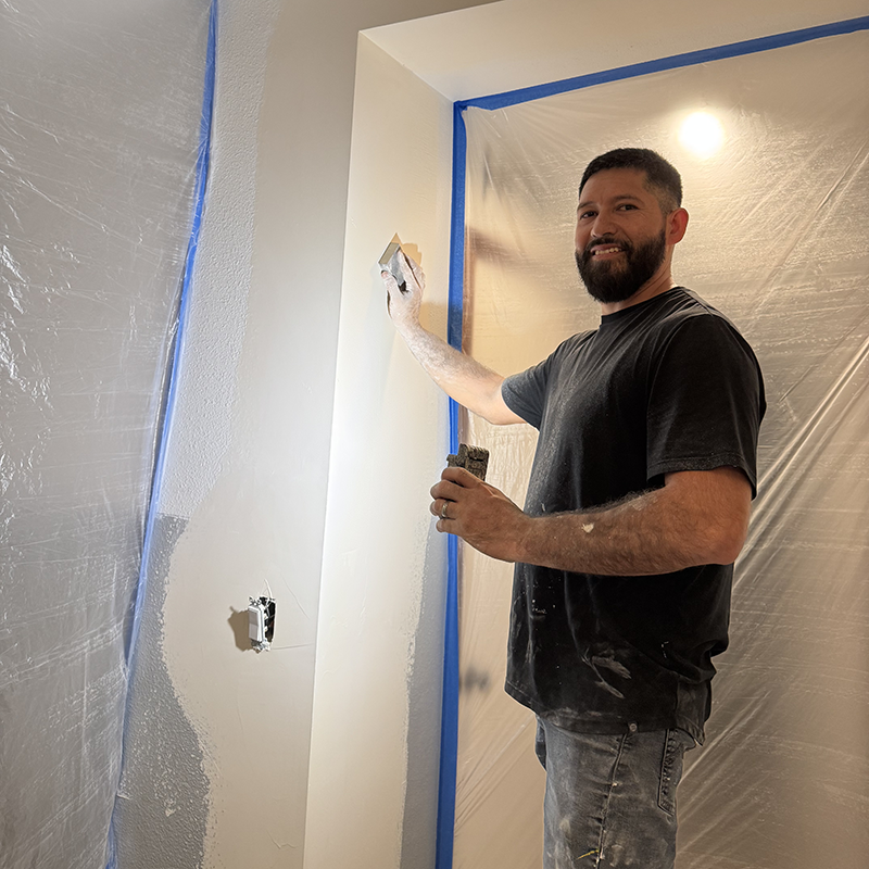 Antonio wearing black shirt and jeans plastering drywall in a room covered with protective plastic sheets.