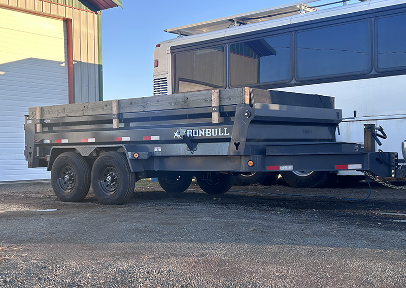 Black Ironbull dual-axle dump trailer parked outside near a white garage door and a large white vehicle.