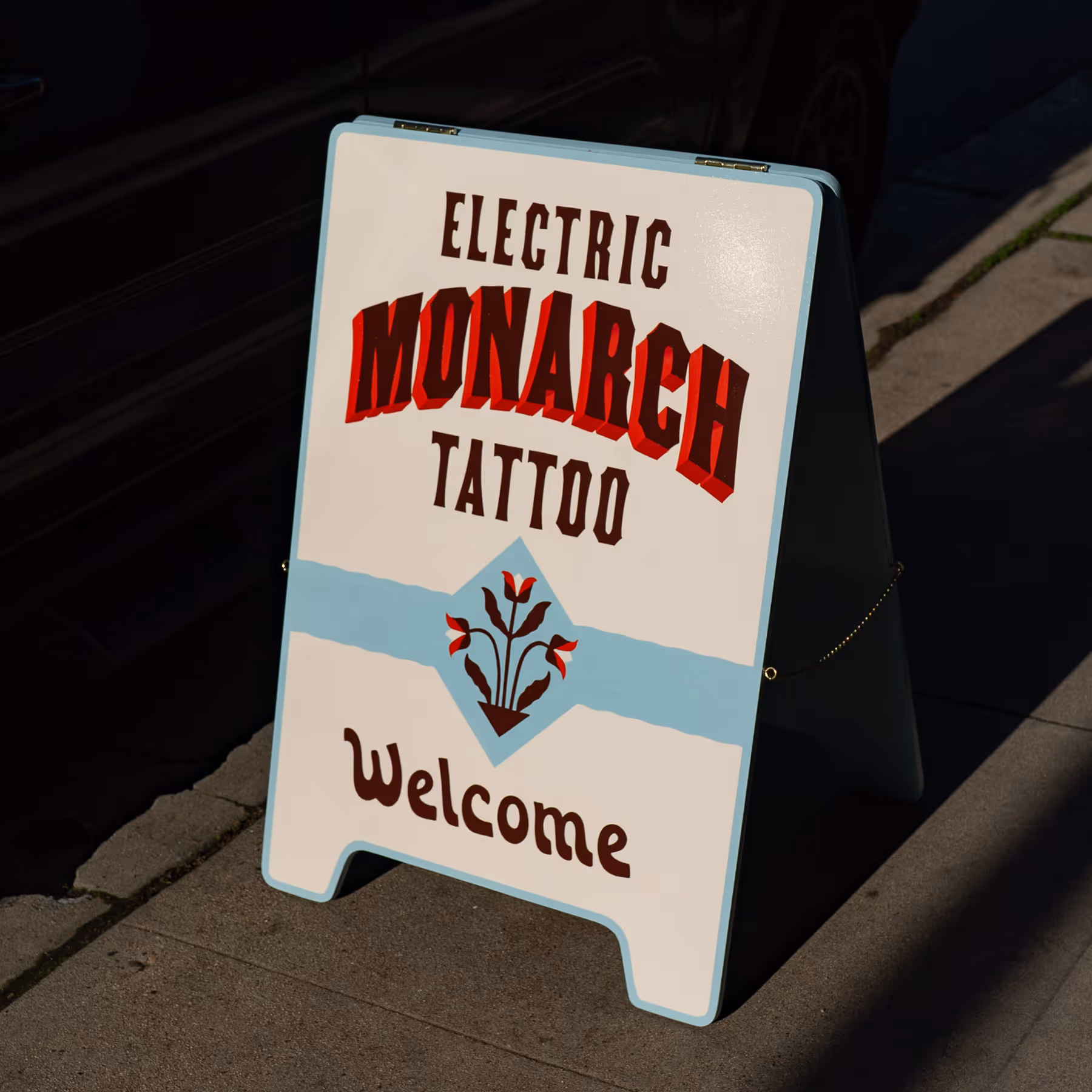 Sidewalk sandwich board sign with text 'Electric Monarch Tattoo Welcome' and a floral design.