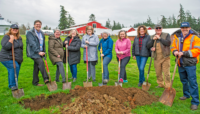 Group of ten people standing in a grassy field holding shovels, posing behind a freshly dug pile of dirt with red barn buildings and trees in the background.