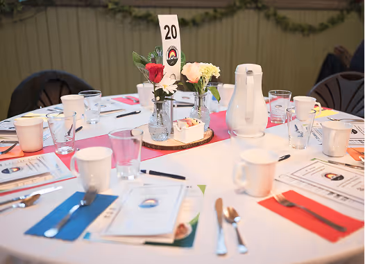 Round dining table set for a meal with white cups, glasses, menus, cutlery on colorful napkins, and floral centerpieces with a table number 20.