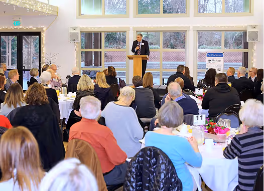 Audience seated at round tables listening to a speaker at a podium in a bright room with large windows.