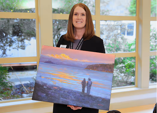 Woman with red hair holding a painting of two figures on a beach at sunset with mountains in the background.