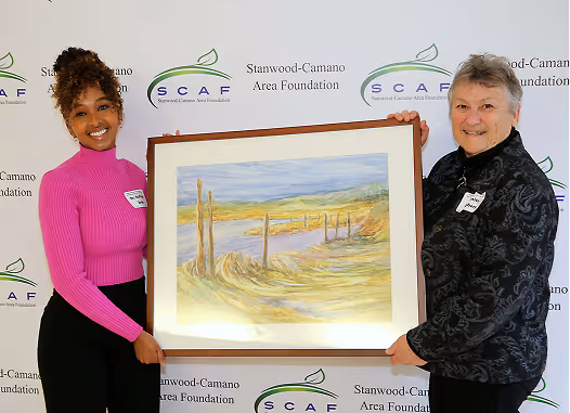 Two women smiling and holding a framed landscape painting with grassy dunes and a water body, standing in front of a Stanwood-Camano Area Foundation banner.