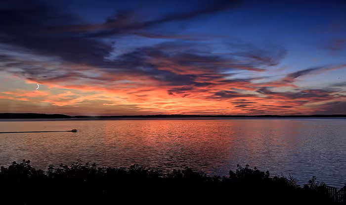 Sunset over a calm lake with a small boat moving across the water and colorful clouds in the sky.