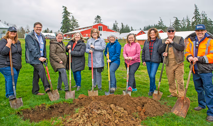 Group of ten people standing in a row on grass, each holding a shovel over a dug hole, with red barns and trees in the background.