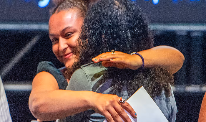 Two women warmly hugging each other, one holding a piece of paper and smiling.