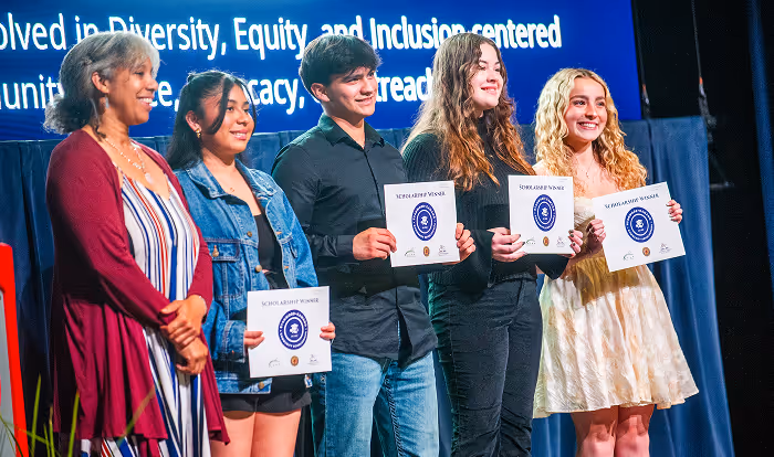 Four young people holding scholarship winner certificates standing next to a smiling woman on stage with blue backdrop text.