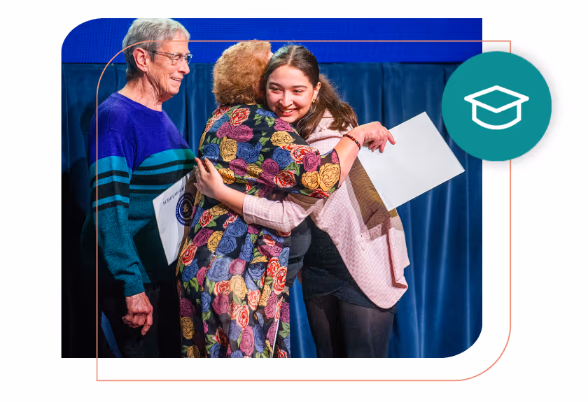 Young woman hugging an older woman while holding a certificate, with an elderly man standing beside them on a stage with blue curtains.