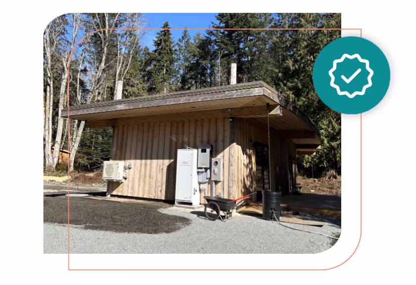Wooden cabin with outdoor equipment including a wheelbarrow and utility boxes, surrounded by trees under a clear blue sky.