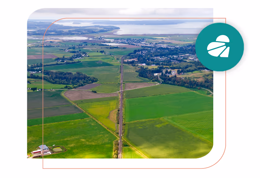 Aerial view of green farmland divided by roads and a railway track, with a body of water in the distance.