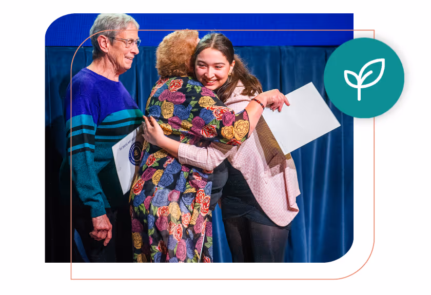 Young woman hugging an older woman on stage while holding a certificate, with another older woman standing nearby also holding a certificate.
