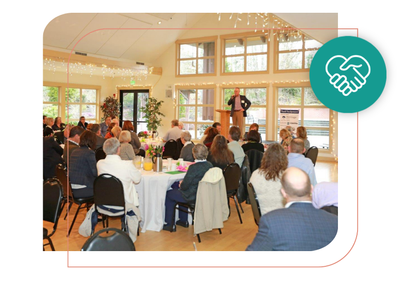 People seated at round tables listening to a speaker at a podium in a room with large windows and string lights.
