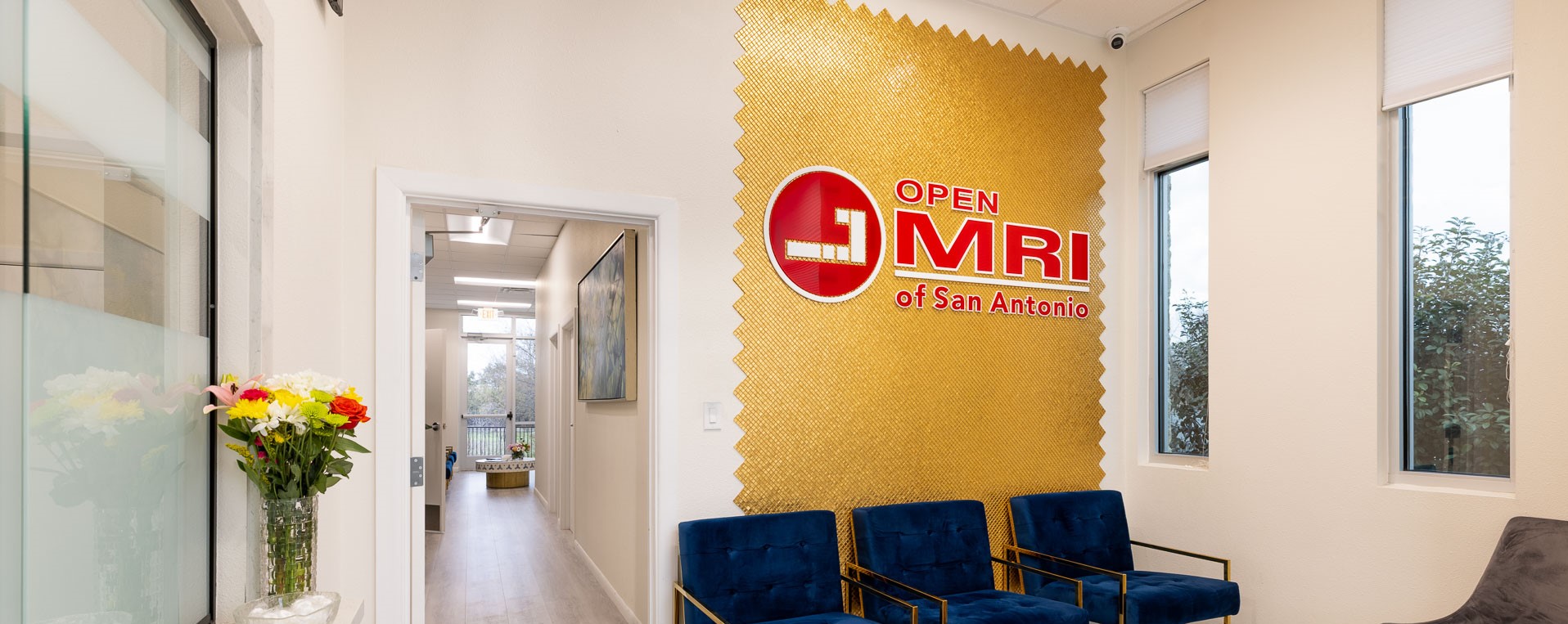 Waiting room with blue chairs, a gold textured wall with Open MRI of San Antonio logo, and windows showing greenery outside.