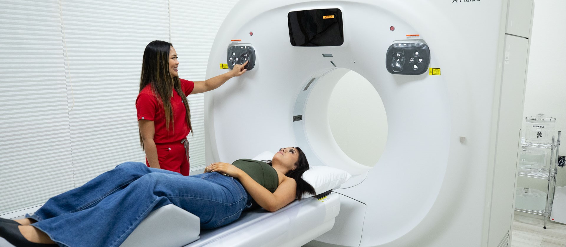 Female technician in red scrubs preparing a woman for a CT scan while the woman lies on the CT scanner table.